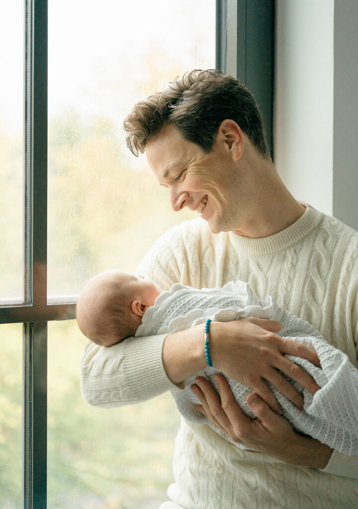 new dad in a cream sweater holds his son by the window of a studio in amsterdam west