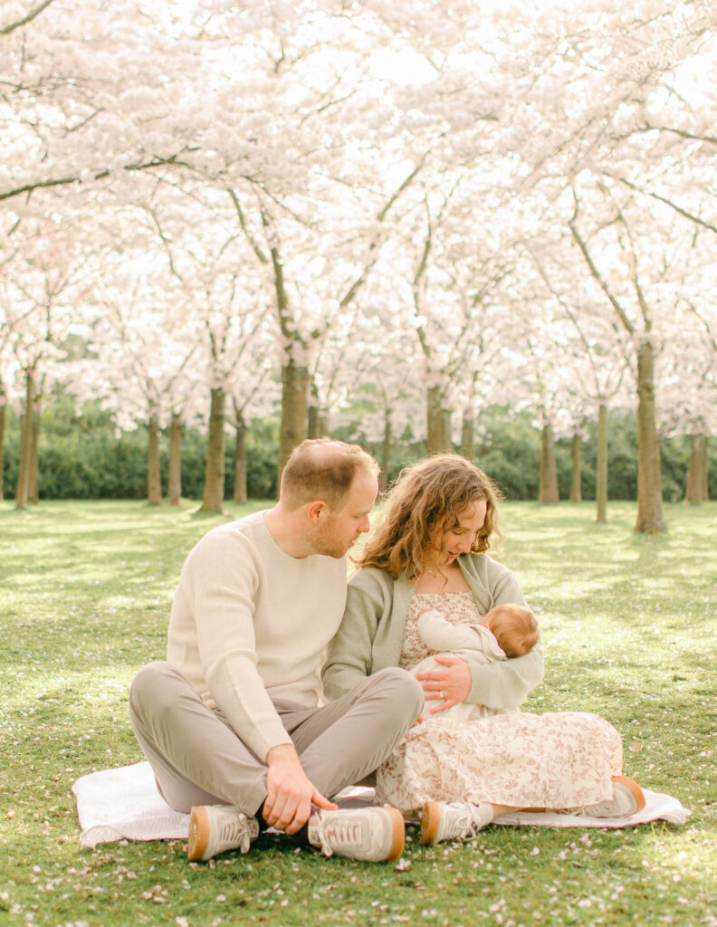 Family of three cuddle on the grass under the cherry blossoms.