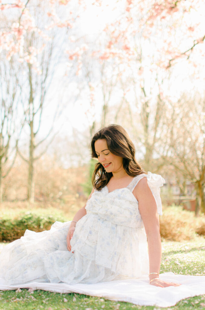 Expecting mother lies on a blanket on the grass under the cherry blossoms.
