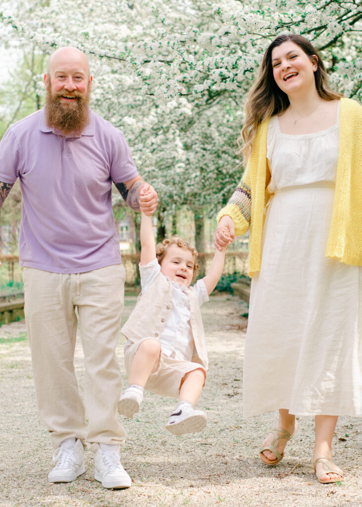 A little boy dressed in white swings happily between his parents in a park.