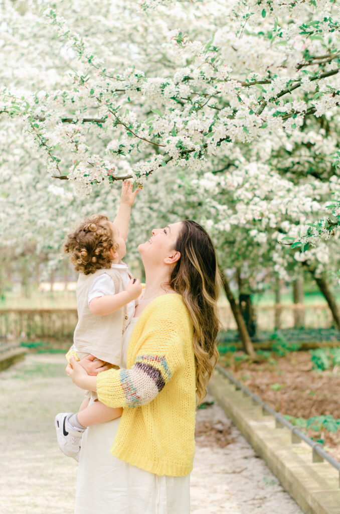 Editorial-style photograph of a mom lifting her son up so he can reach the branches of an almond blossom tree.