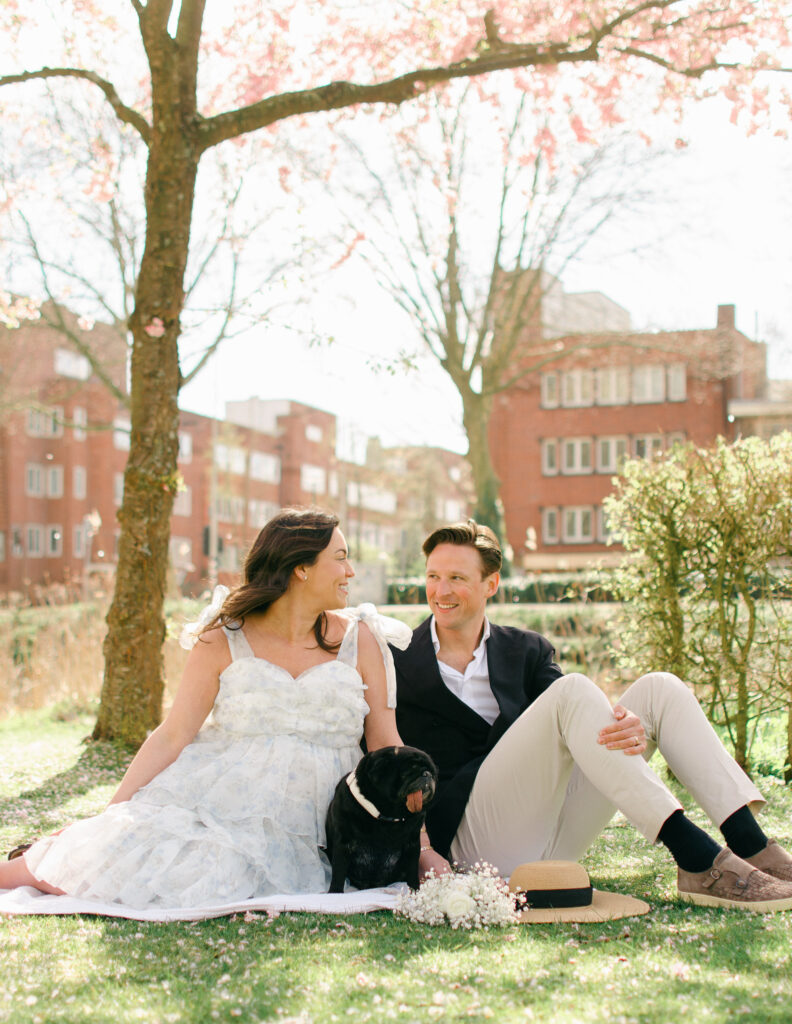Pregnant couple sits on a white blanket under the cherry blossoms in Erasmuspark.