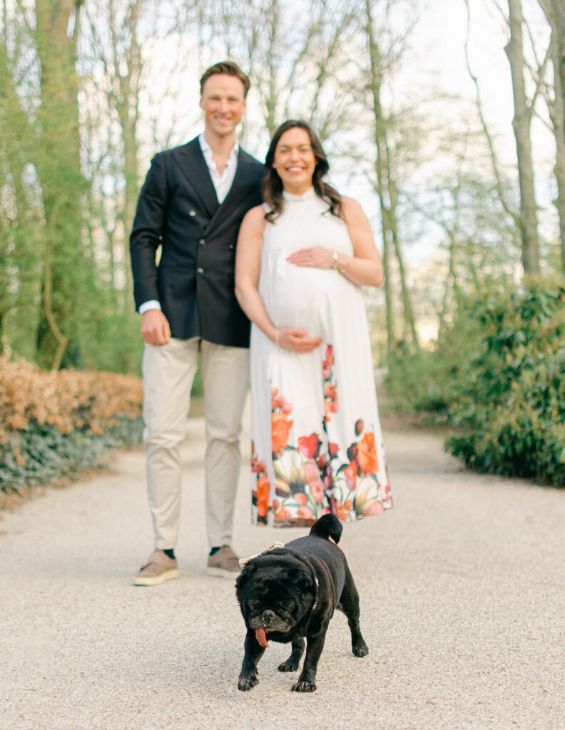 "Portrait of expecting couple with arms around each other, dog on leash beside them"