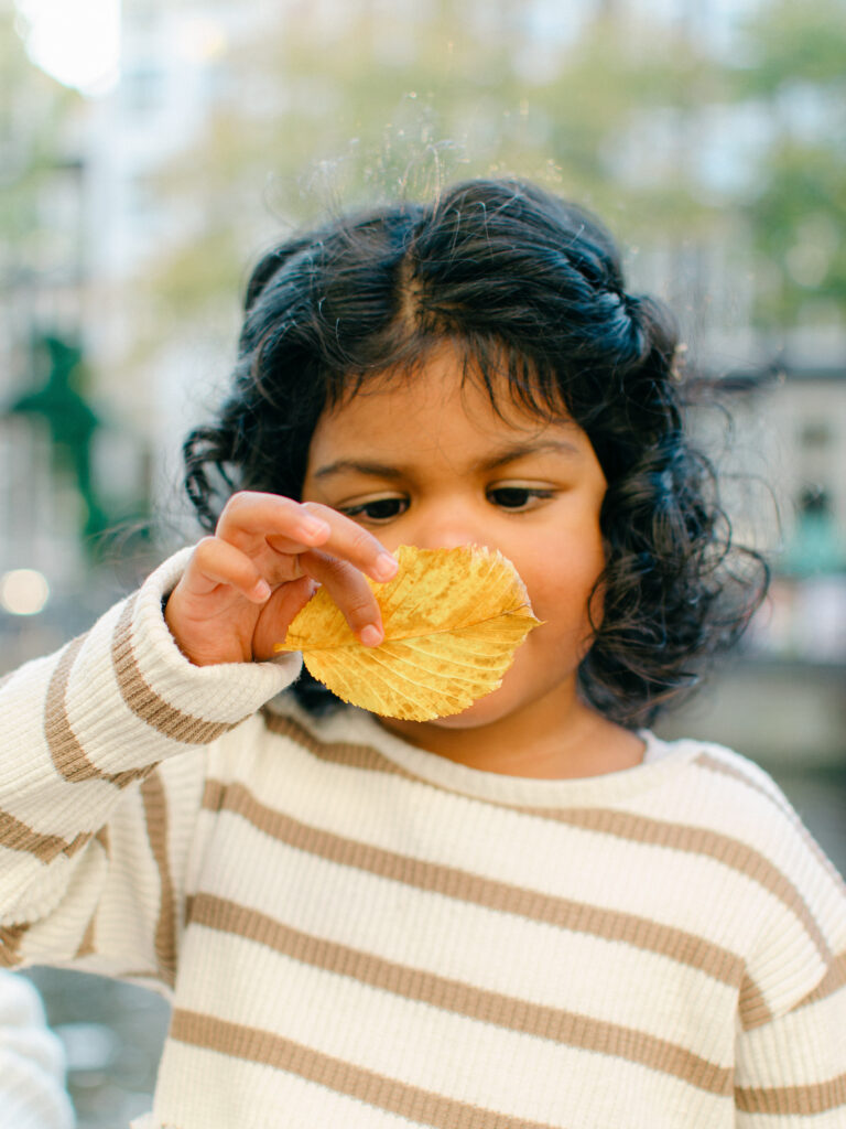 A little girl holds a yellow leaf up to inspect it in Amsterdam in fall. She is wearing neutrals and has curly dark hair.