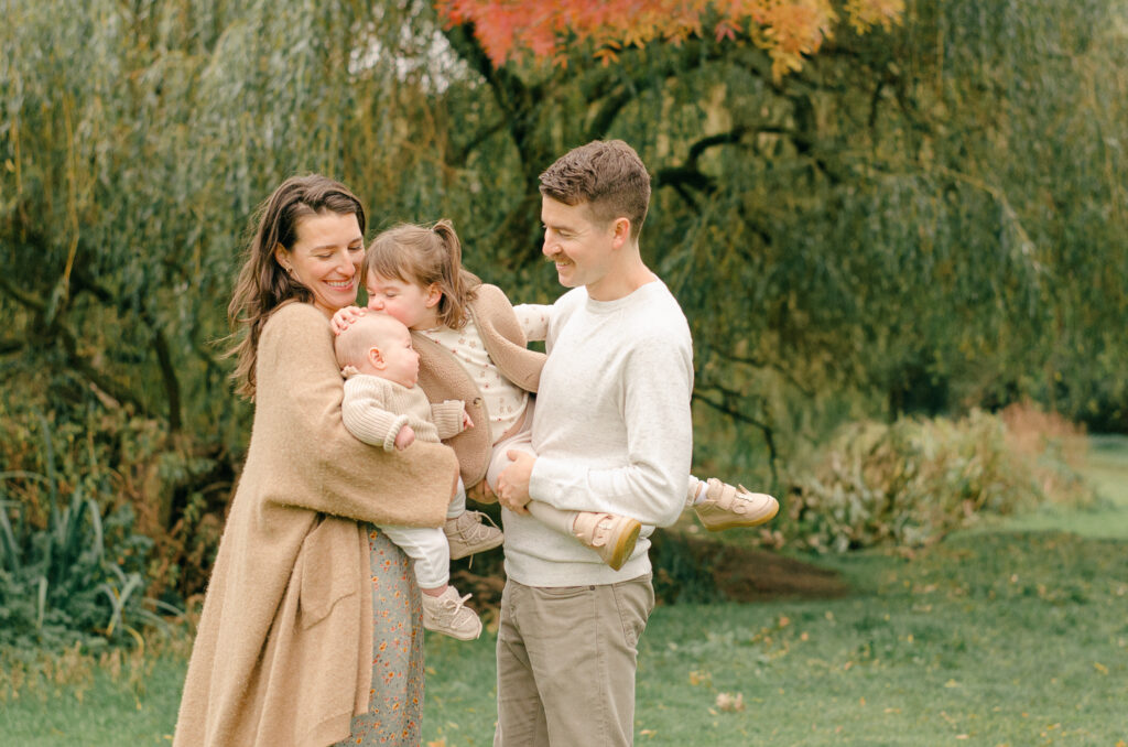 Parents hold daughters up so the toddler can kiss the baby on the top of the head.