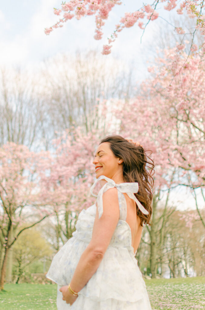 "Side profile of pregnant woman in floral dress holding belly under spring blossoms"
