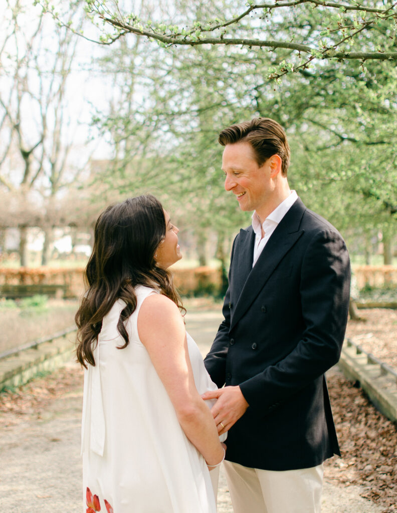 Backlit photo of couple embracing while mother cradles her bump, while the light sprinkles through the trees.