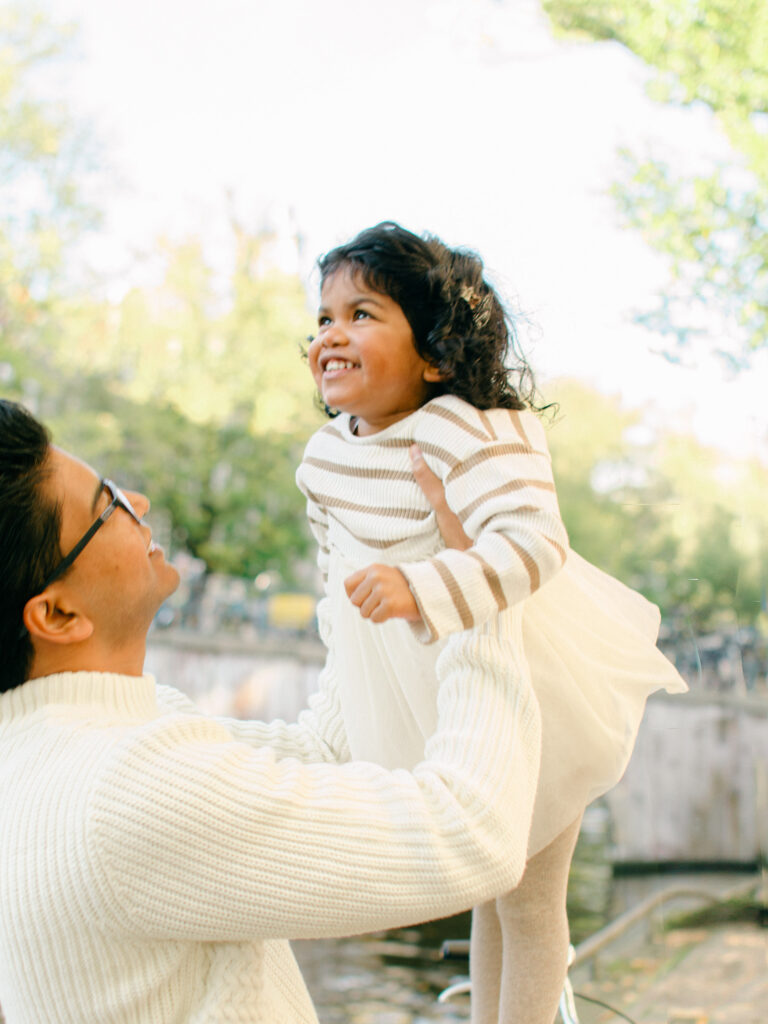 Bright, fun portrait of dad lifting young daughter up into the air with the fall leaves behind them.
