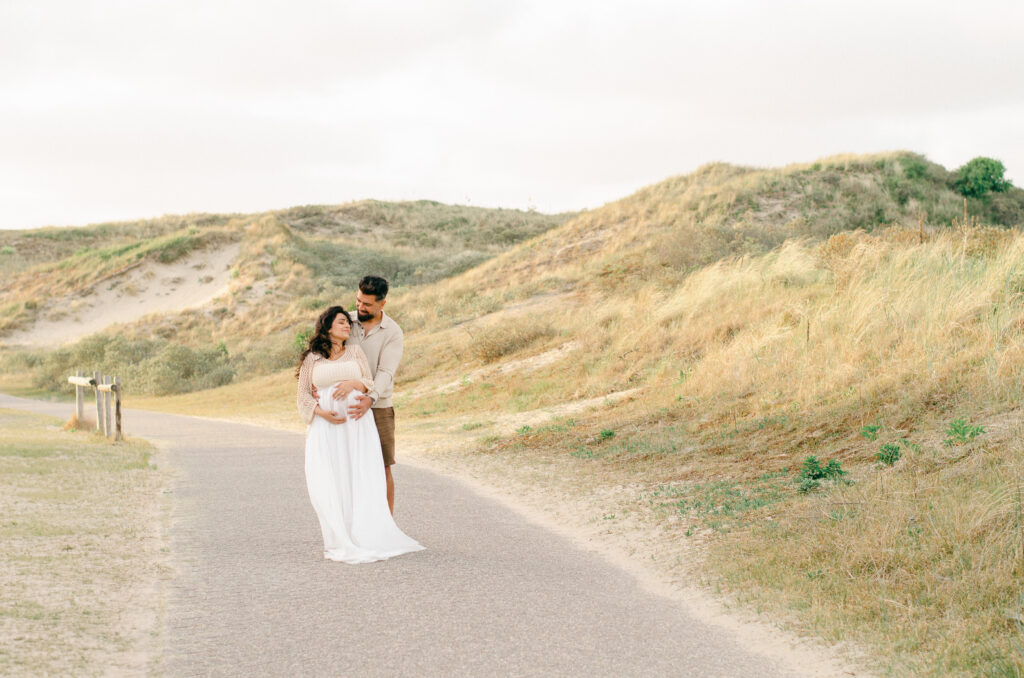 A horizontal image of a husband and wife embracing in the Bloemendaal dunes with rolling hills drenched in sunshine behind them.