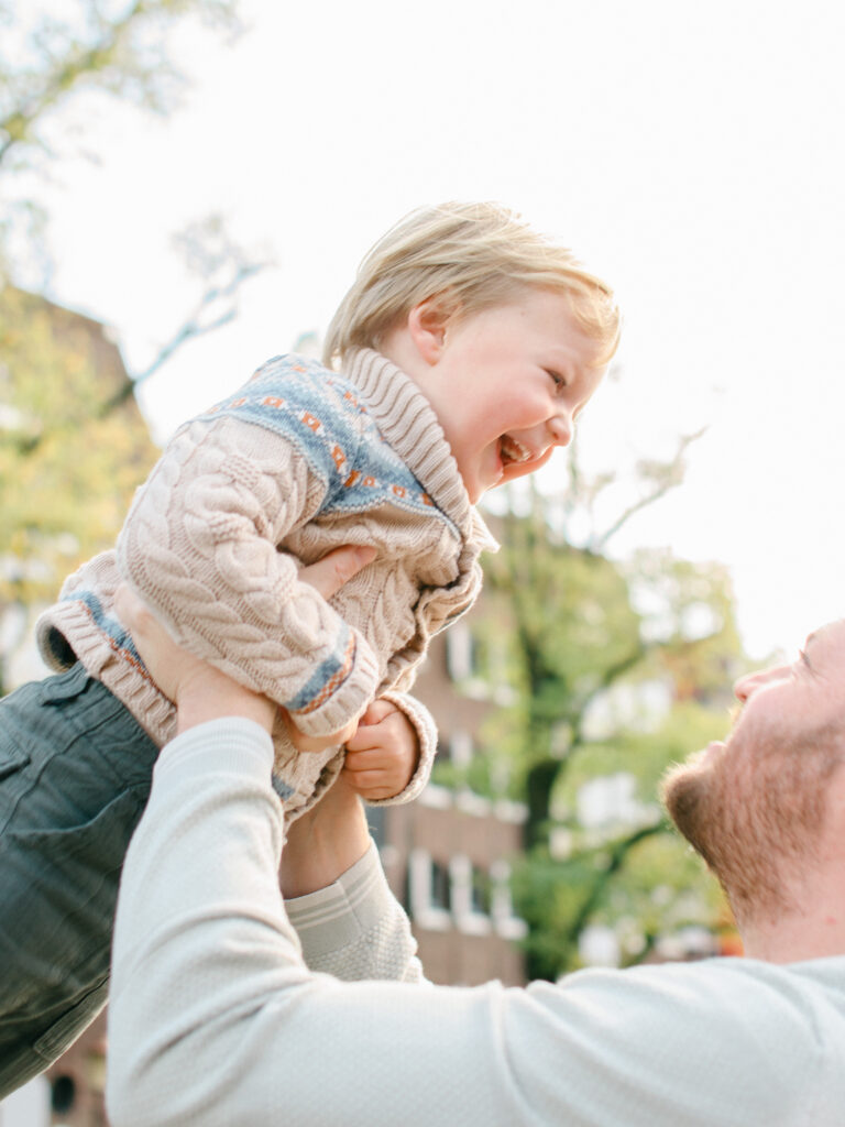 Dad lifts laughing boy into the air, with sunlight in the background.