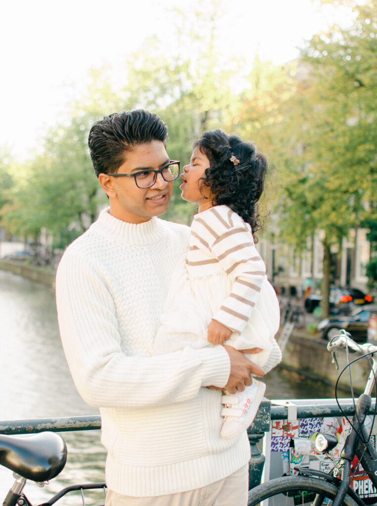 Toddler leans in to give her Papa a kiss on the cheek in Amsterdam in the fall. They are wearing neutrals.