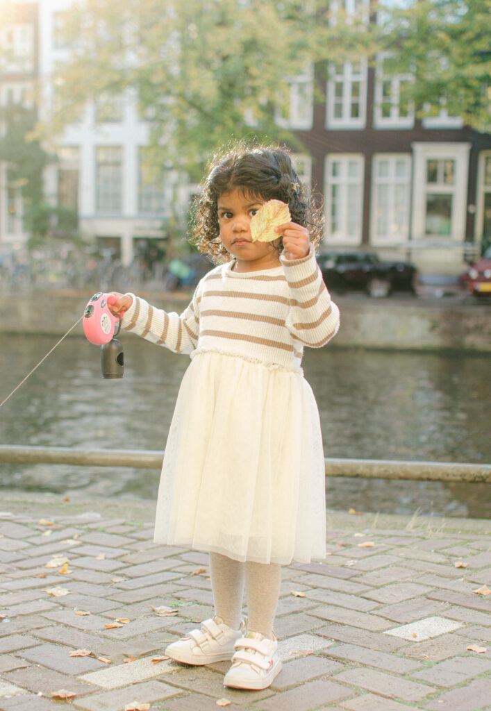 Toddler plays in the fall leaves on the Keizersgracht canal and holds her dog's leash.