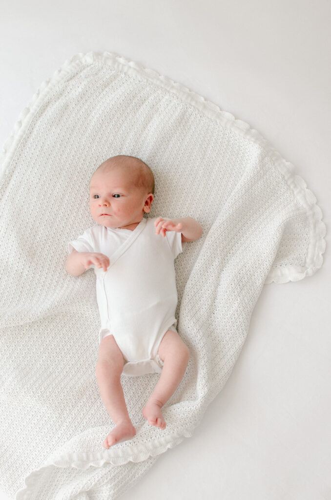 Baby sprawled out on a white blanket on a white backdrop, wide awake.