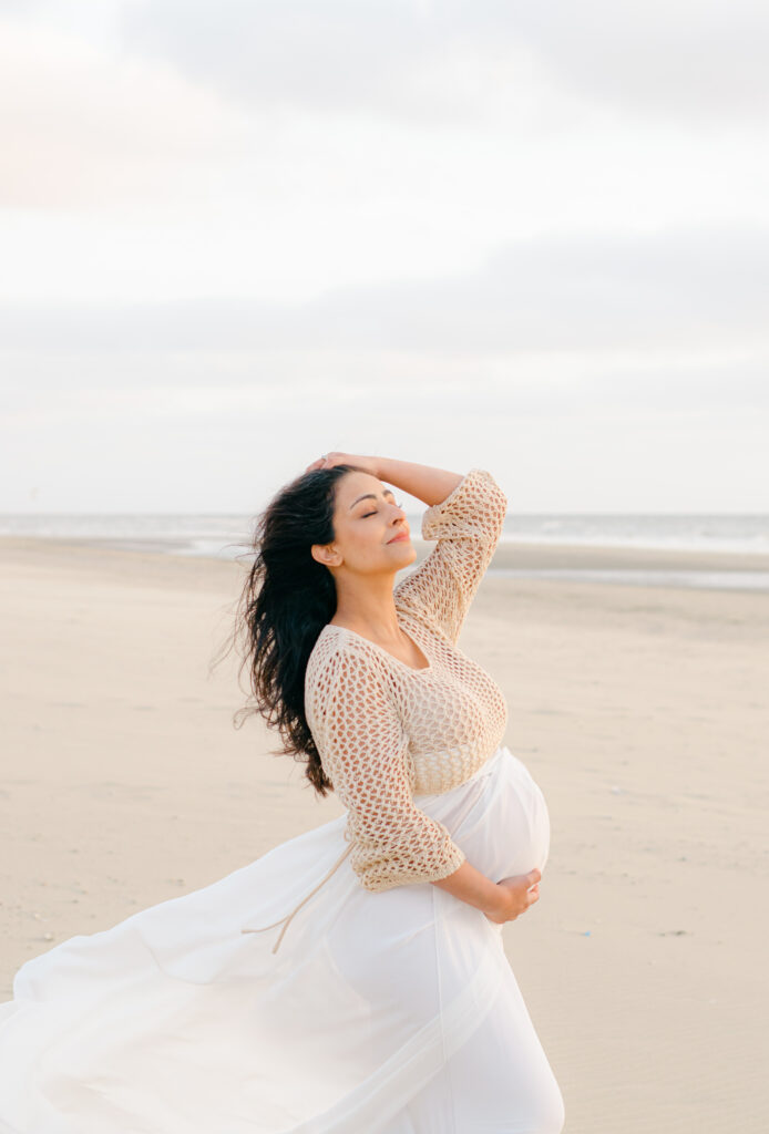 A pregnant mama brushes her hair back against the wind and holds her belly with her other hand in Bloemendaal.