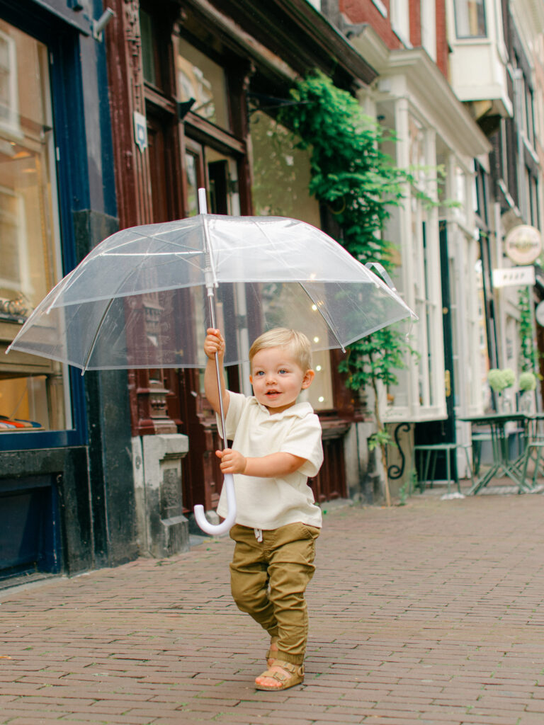 Toddler boy wearing white and khaki runs down the cobblestone streets of Amsterdam holding a clear umbrella.