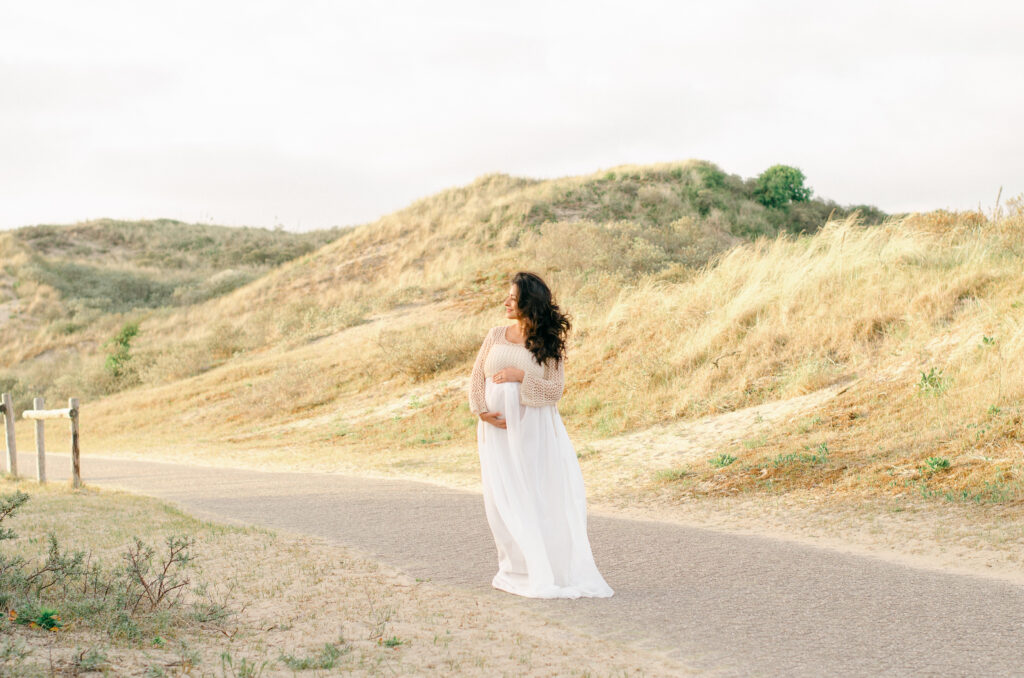 A pregnant woman wearing a flowing white dress cradles her stomach with the sunsetting on the dunes behind her.