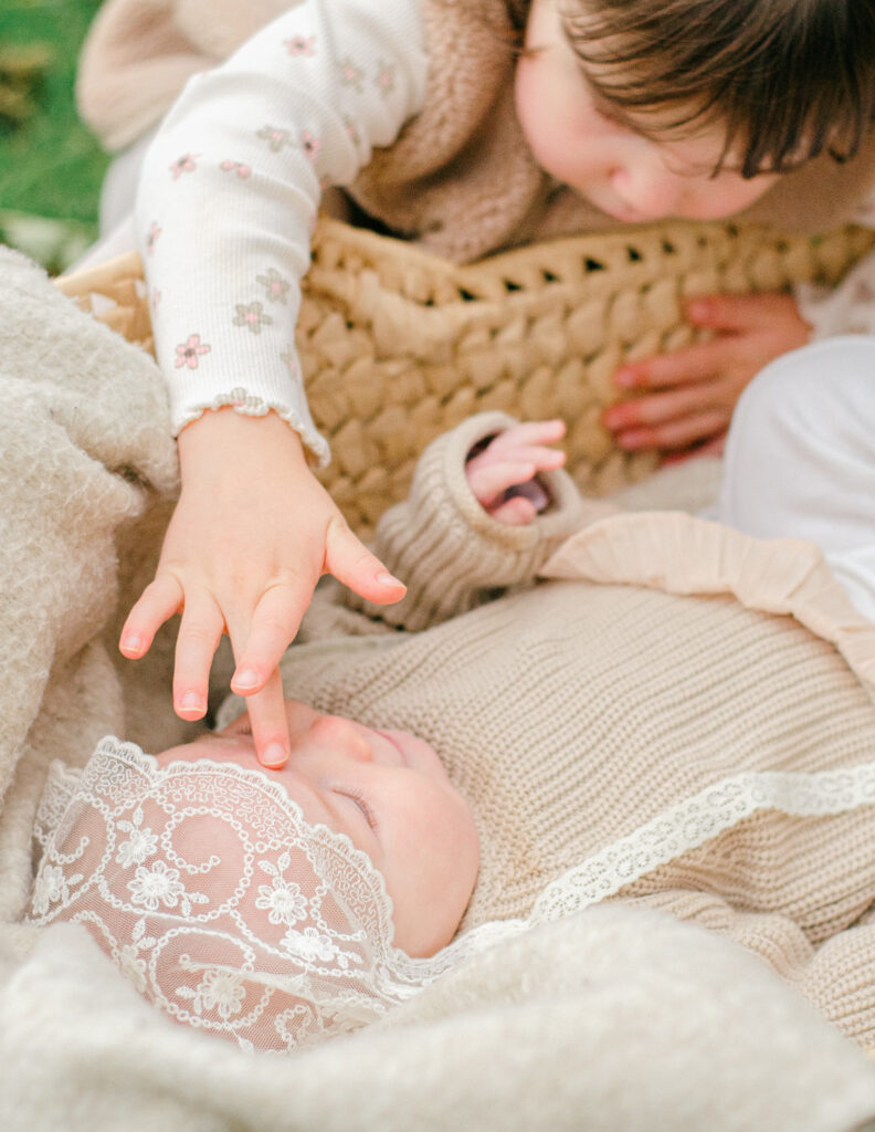 Toddler teaches out to touch little sister's face while she lies down in the bassinet.