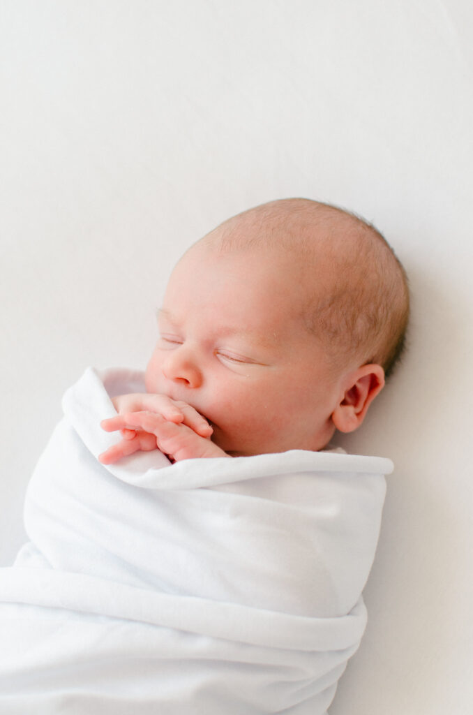 Baby wrapped in a white fabric, his hands tucked under his chin, resting peacefully.