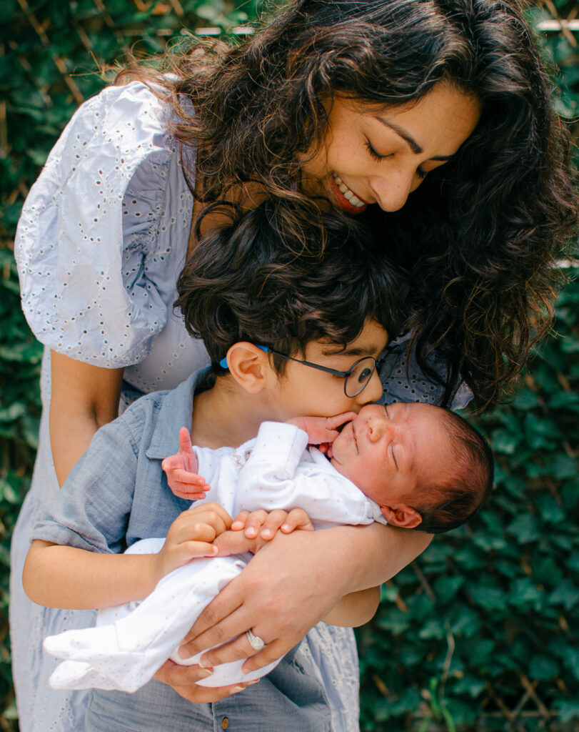 Mom holds baby so toddler can give him a big kiss. 