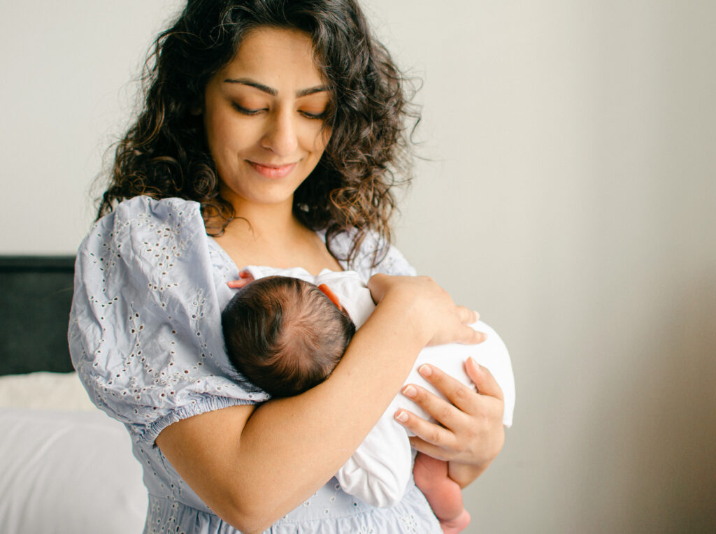 Mother snuggles newborn baby near the window in her bedroom during a lifestyle home shoot.