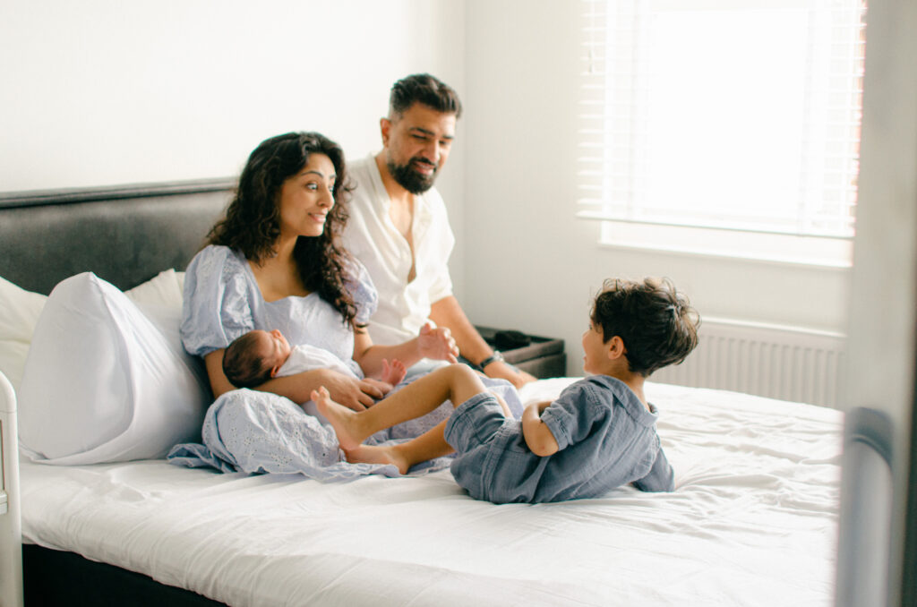 Toddler jumps on bed while his parents laugh during a newborn shoot.