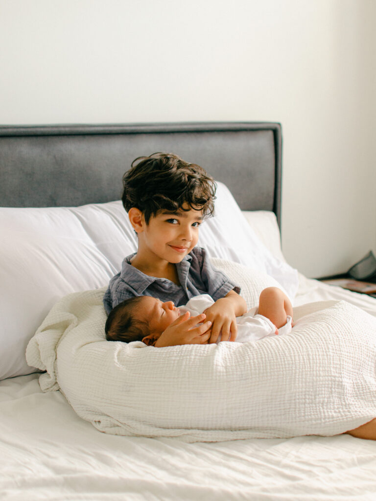 Older brother sits and holds his newborn baby brother on the bed.