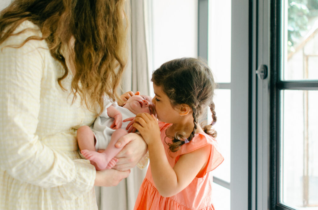 Little sister kisses her baby sister during a newborn session in their home. 