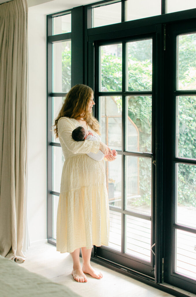Mother looks out the window towards the garden during her lifestyle home newborn session in Oud-West.