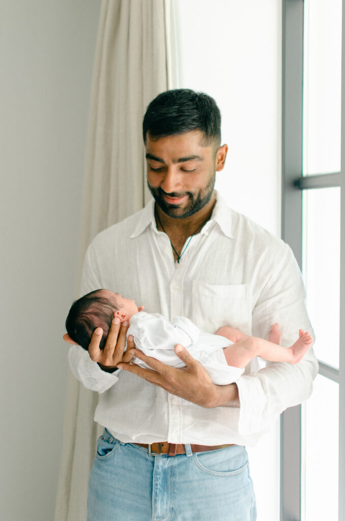Dad holds newborn baby in front of the window.
