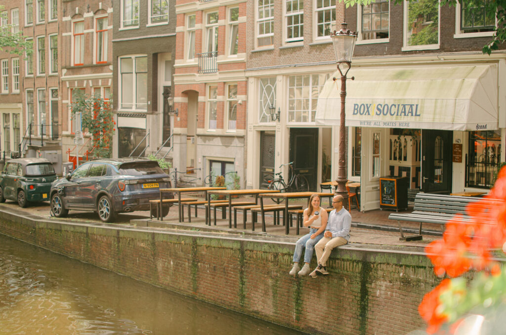 A couple sits on the edge of the canal, dangling their legs and drinking coffee in the summer. 